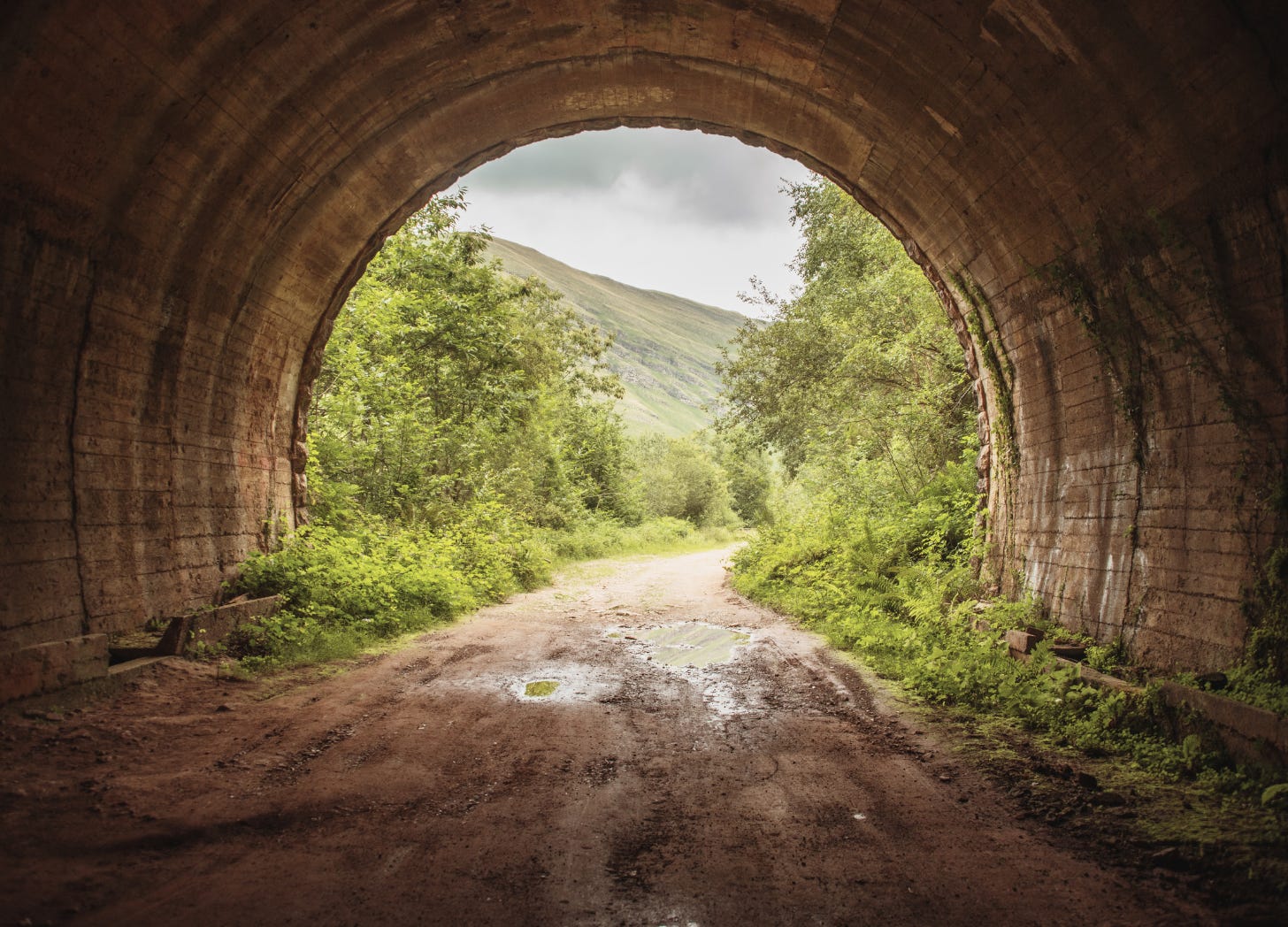 View from inside a tunnel looking out onto a dirt path, with trees and hills visible in the light beyond.