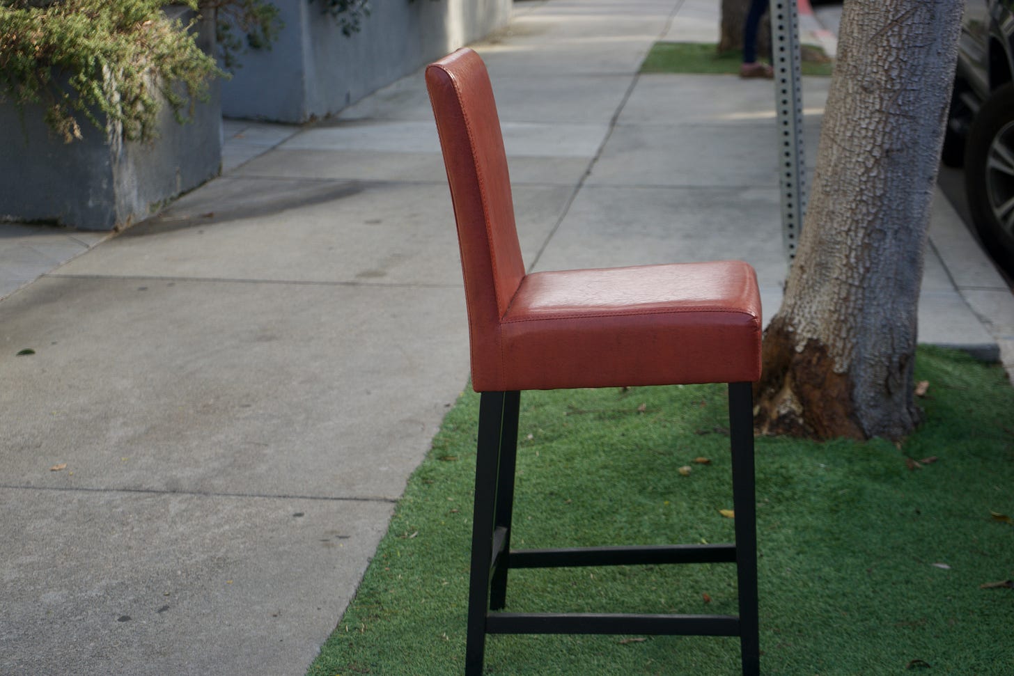 Side-profile of a rust-toned leather barstool placed precisely on a narrow strip of artificial grass between sidewalk and curb, framed by clean horizontal concrete lines and a tree trunk, with generous negative space emphasizing isolation and intention. Side-profile of a rust-toned leather barstool placed precisely on a narrow strip of artificial grass between sidewalk and curb, framed by clean horizontal concrete lines and a tree trunk, with generous negative space emphasizing isolation and intention.