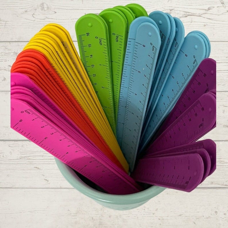 May include: A collection of colorful rulers in a variety of shades, including pink, orange, yellow, green, blue, and purple. Each ruler is approximately 6 inches long and is displayed in a light blue bowl against a white wood background.