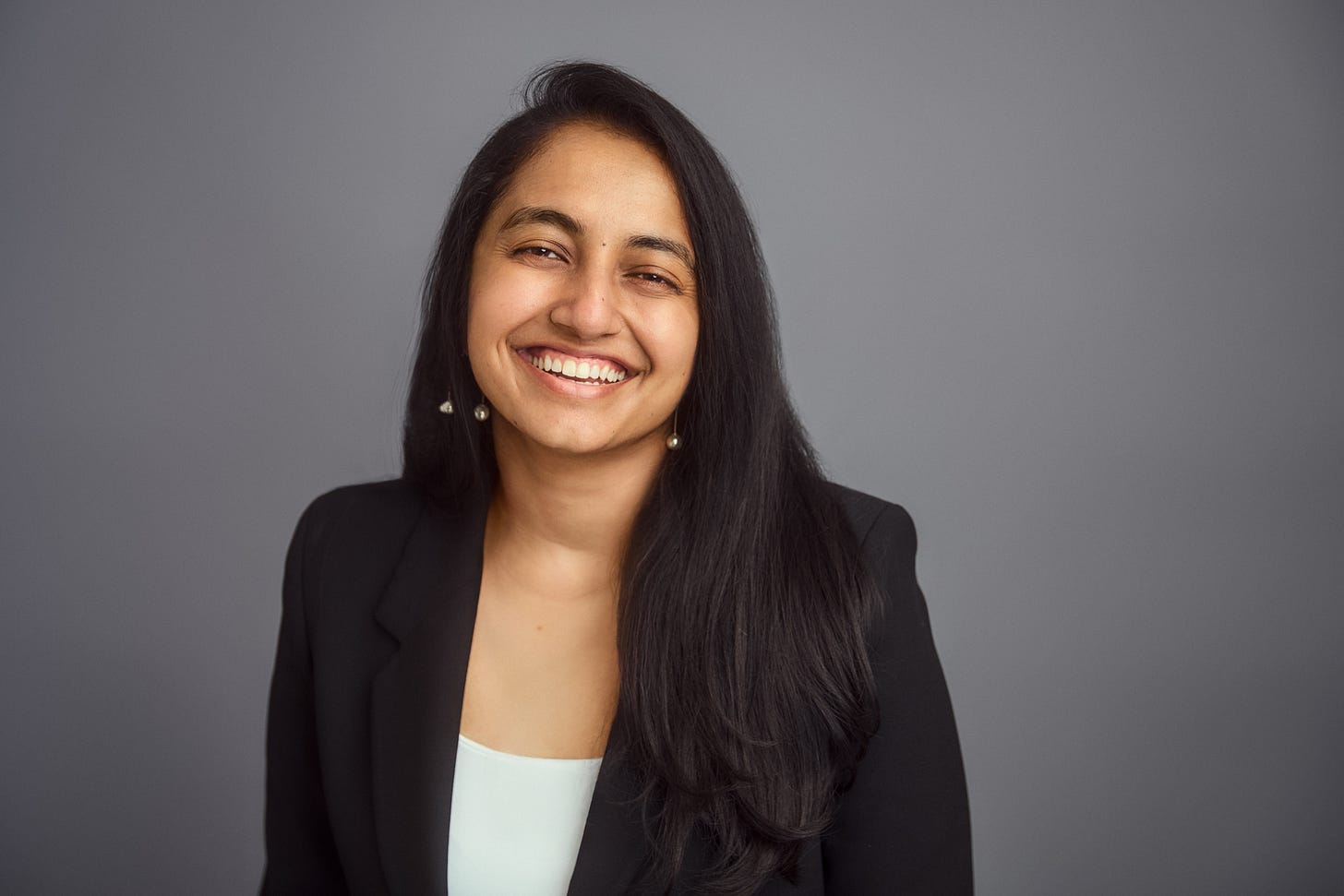 A brown person with black hair falling past their shoulders looks at the camera, beginning to smile. They wear a black blazer over a white shirt. A brown person with black hair falling past their shoulders looks at the camera, beginning to smile. They wear a black blazer over a white shirt.