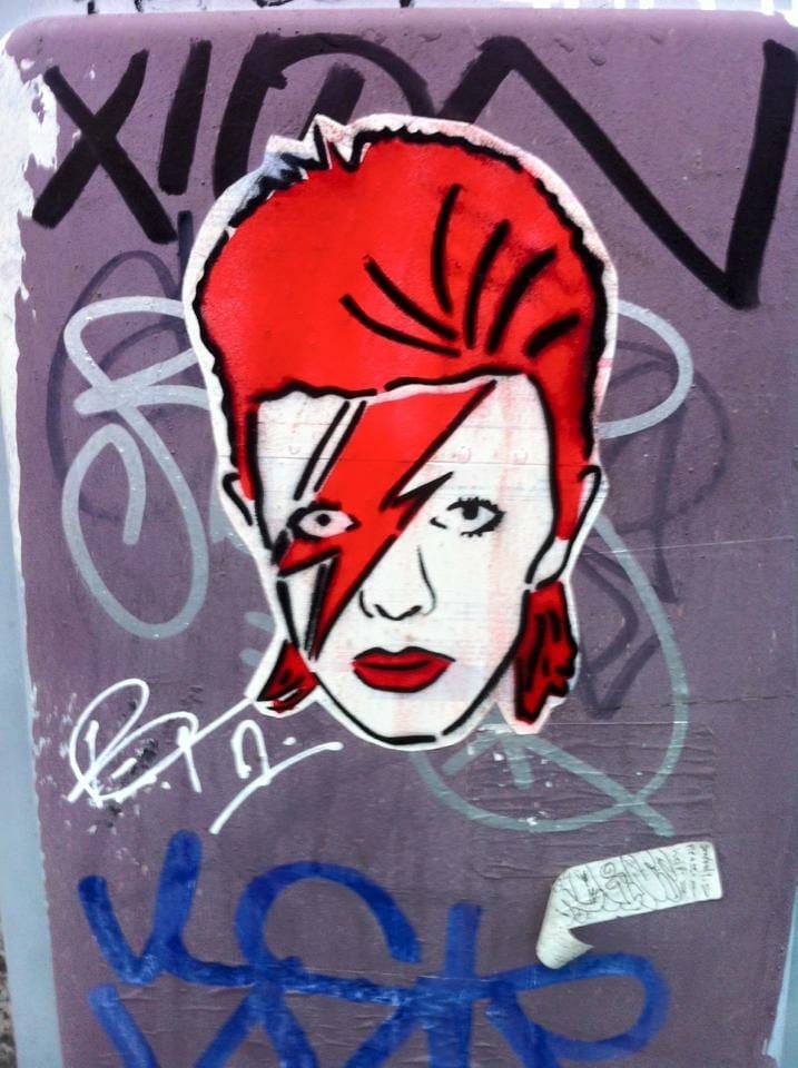 Graffiti of a stylized face with red hair and a bold red lightning bolt across the face on a purple, graffiti-covered wall. Energetic and rebellious tone. The portrait is of David Bowie. He has a tall red mullet, and a red lightning bolt across his face, and has red lipstick on. Graffiti of a stylized face with red hair and a bold red lightning bolt across the face on a purple, graffiti-covered wall. Energetic and rebellious tone. The portrait is of David Bowie. He has a tall red mullet, and a red lightning bolt across his face, and has red lipstick on.