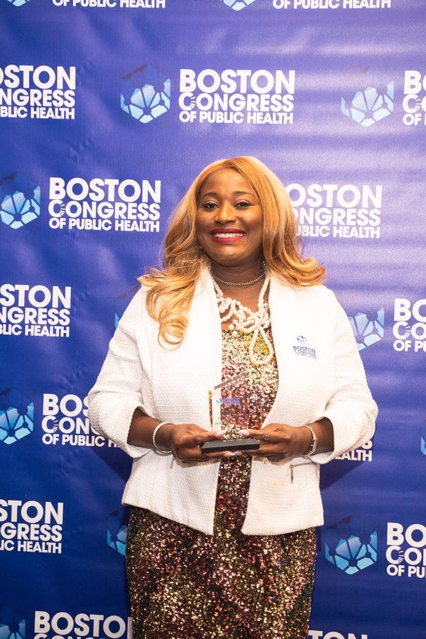 Dr. Oluwatosin Olateju in a white blazer over a sequined dress, smiling with her award on the BCPH backdrop.