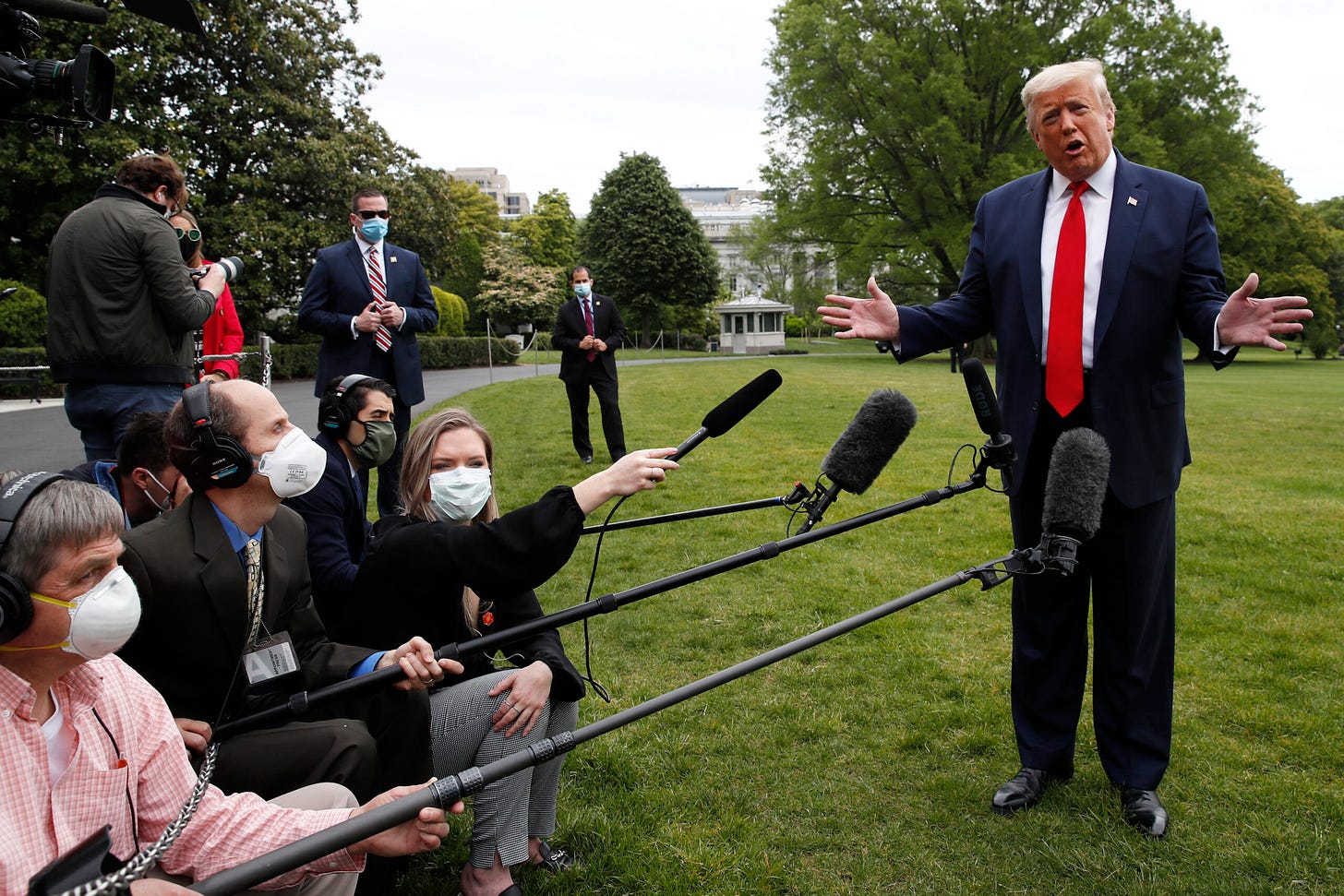 Donald Trump is standing outside on a lawn, with reporters using long mics to interview him. The reporters are wearing face masks and are kneeling down.