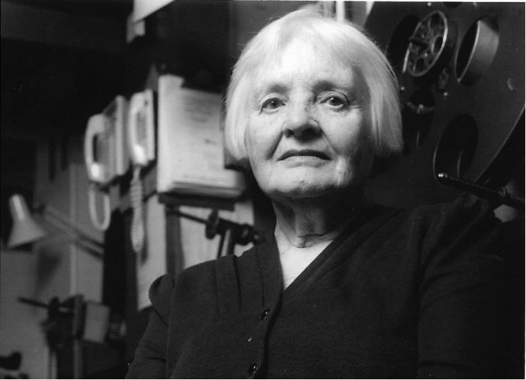 A black and white photo of an older woman with short white hair wearing dark clothes. There are multiple telephones and a lamp in the background.