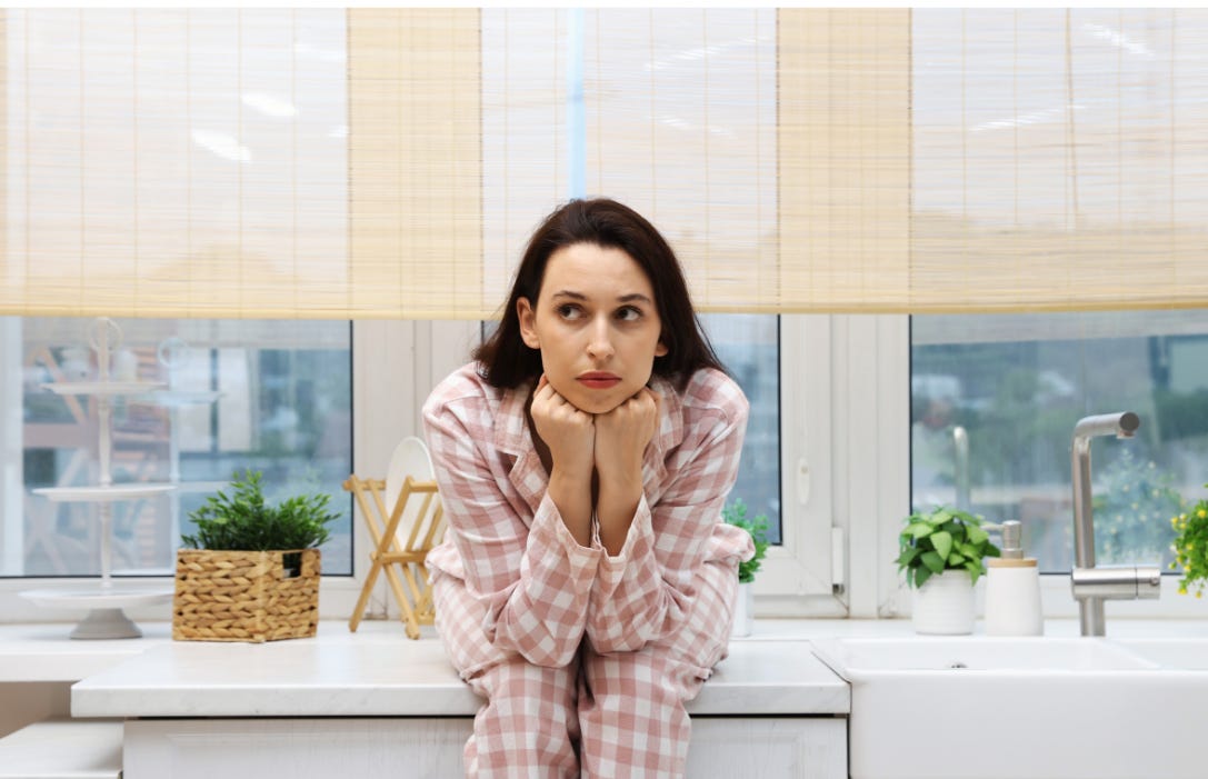 A woman in plaid pajamas sits on her kitchen counter, chin resting on her fists, looking weary and paused in a bright, neutral kitchen.
