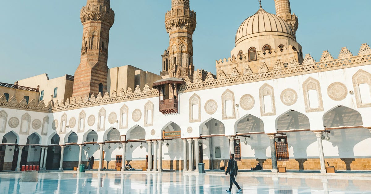 A man walks through the courtyard of Al Azhar Mosque in Cairo, surrounded by white columns, minarets and a large dome.