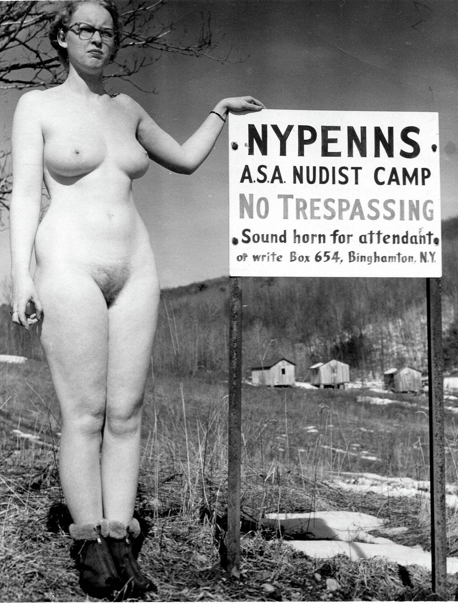A nude woman wearing boots stands beside a sign reading “NYPENNS A.S.A. Nudist Camp — No Trespassing,” with small cabins visible on the hillside behind her.