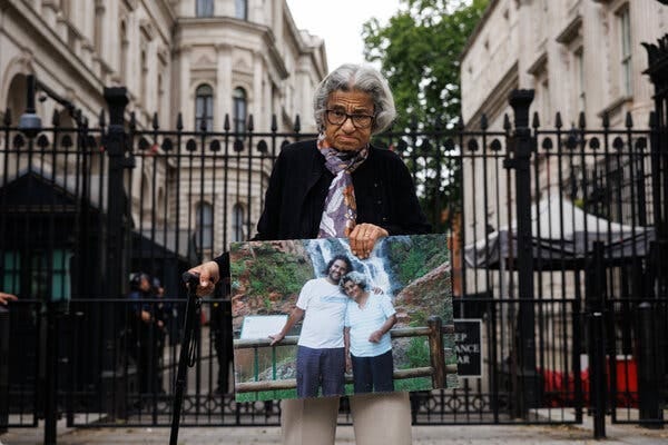 A woman with white hair and a walking stick, standing in front of a gate and holding a large photograph of herself with her son.
