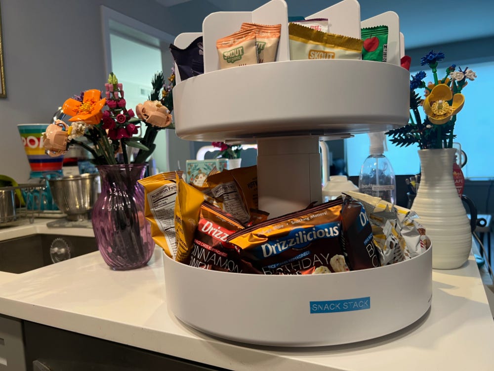 A white plastic two-tiered snack organizer atop a white kitchen counter. The organizer is filled with packages of snacks. On either side is a vase filled with LEGO flowers.