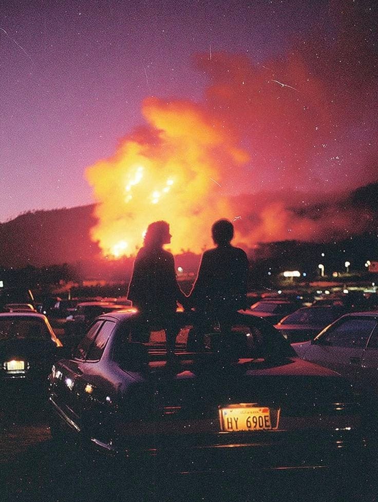 This may contain: two people standing on the hood of a car in front of a fire that is burning This may contain: two people standing on the hood of a car in front of a fire that is burning