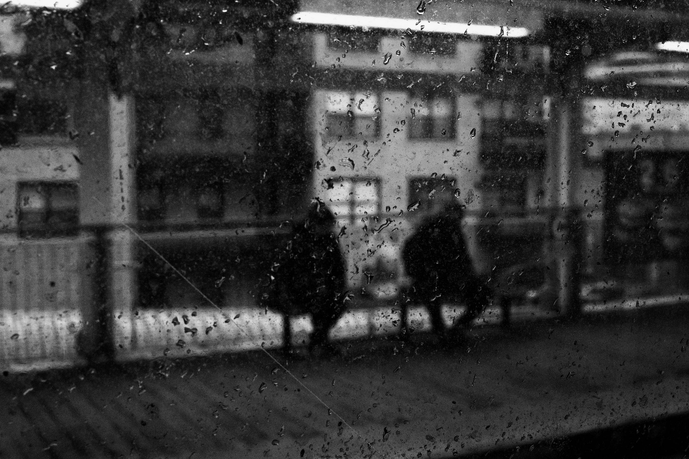 Rain-speckled window blurring the view of two people sitting on a bench at a train station, with buildings and platform lights faintly visible in the background. Rain-speckled window blurring the view of two people sitting on a bench at a train station, with buildings and platform lights faintly visible in the background.