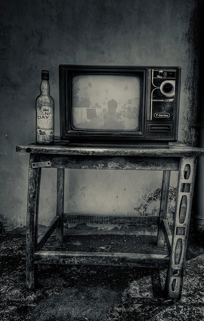 An old television set on top of a dilapidated wooden stand. An empty bottle of booze sits next to the set.