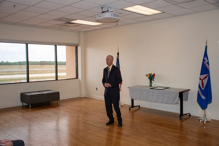 Two men stand in a plain ceremony room with wood floors, holding a framed certificate together. The man on the left wears a white Civil Air Patrol–style uniform; the man on the right wears a dark short-sleeve shirt and khaki pants. The certificate reads “Amelia Earhart Award.” An American flag is on the left and a blue Civil Air Patrol flag (with the red propeller emblem and “PATROL,” plus a “Tallahassee” streamer) is on the right. A small table with a white cloth sits behind them under fluorescent ceiling lights. Also, A man in a dark suit stands speaking in a bright room with hardwood floors and large windows overlooking an airfield. Behind him are a U.S. flag and a blue Civil Air Patrol flag. To the right is a table draped in a cloth with a framed certificate and a vase of flowers. An Epson ceiling-mounted projector hangs above, and a small bench sits by the windows.