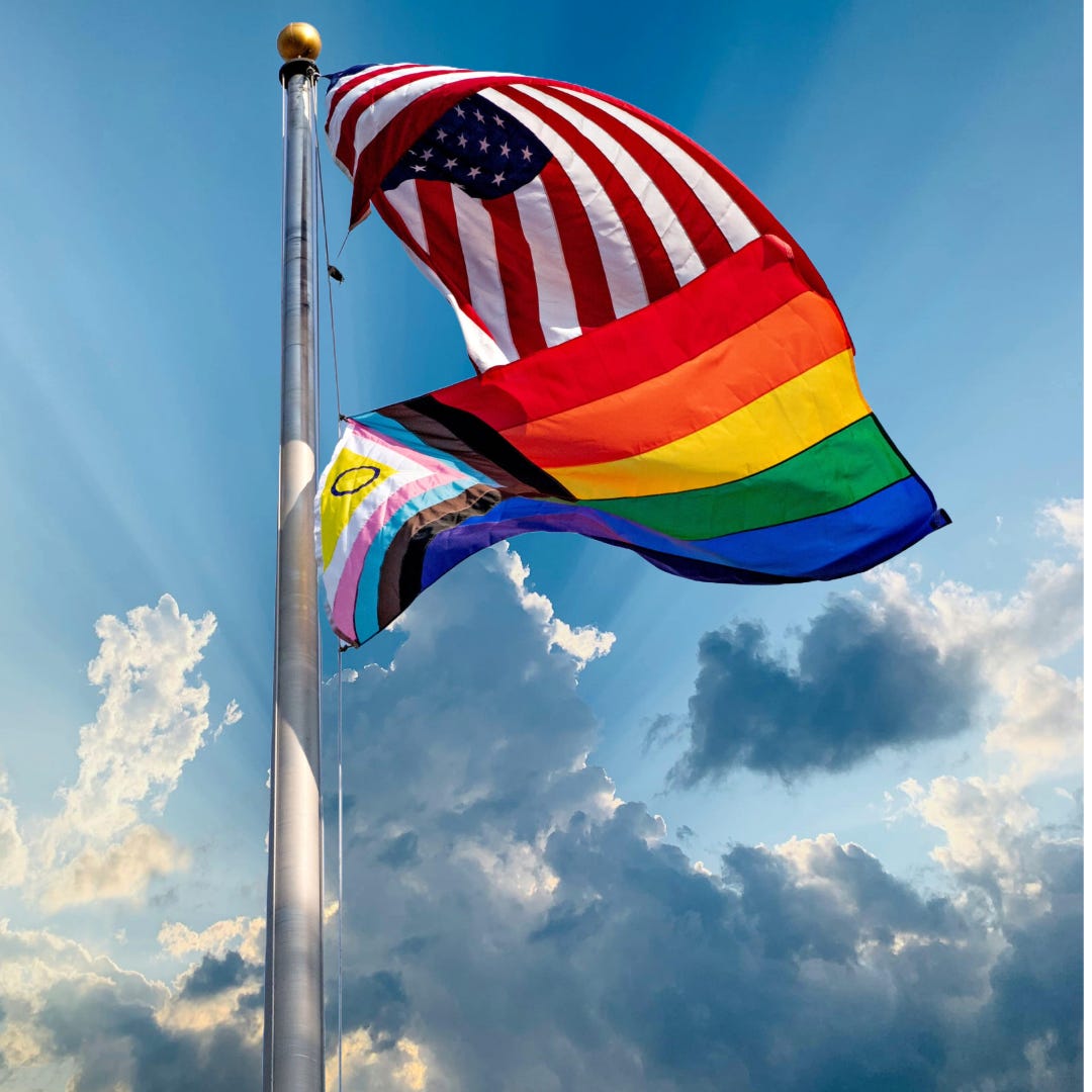 An American flag and an inclusive Pride flag against a blue sky with clouds.