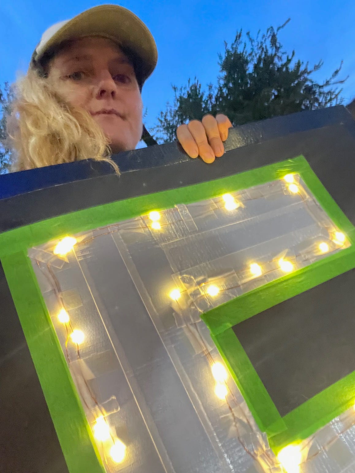 A white woman in a baseball cap holds up a sign - we can see it is a large E in twinkle lights