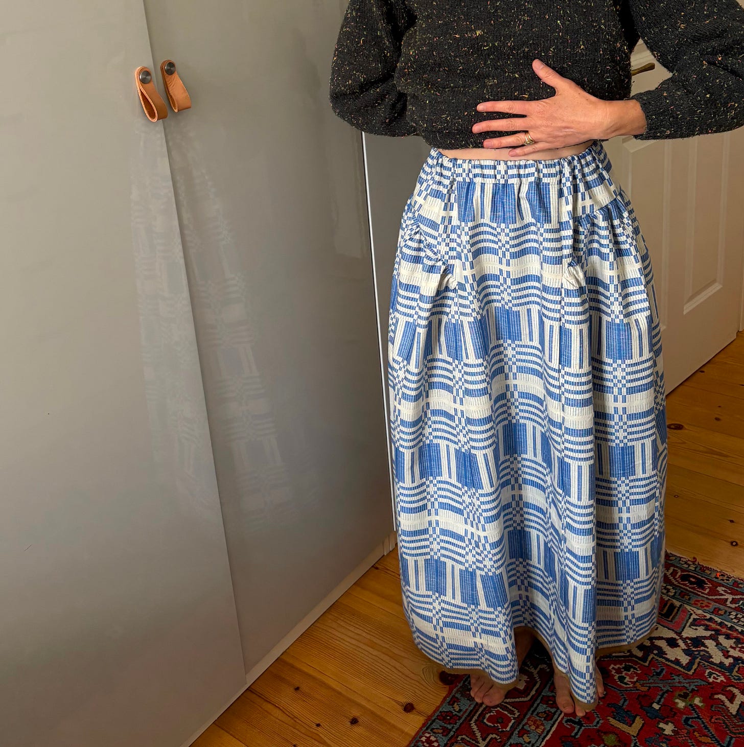 A woman in a dark sweater and blue-and-white patterned skirt stands barefoot on a Turkish carpet and wood floor next to a grey wardrobe door. A woman in a dark sweater and blue-and-white patterned skirt stands barefoot on a Turkish carpet and wood floor next to a grey wardrobe door.
