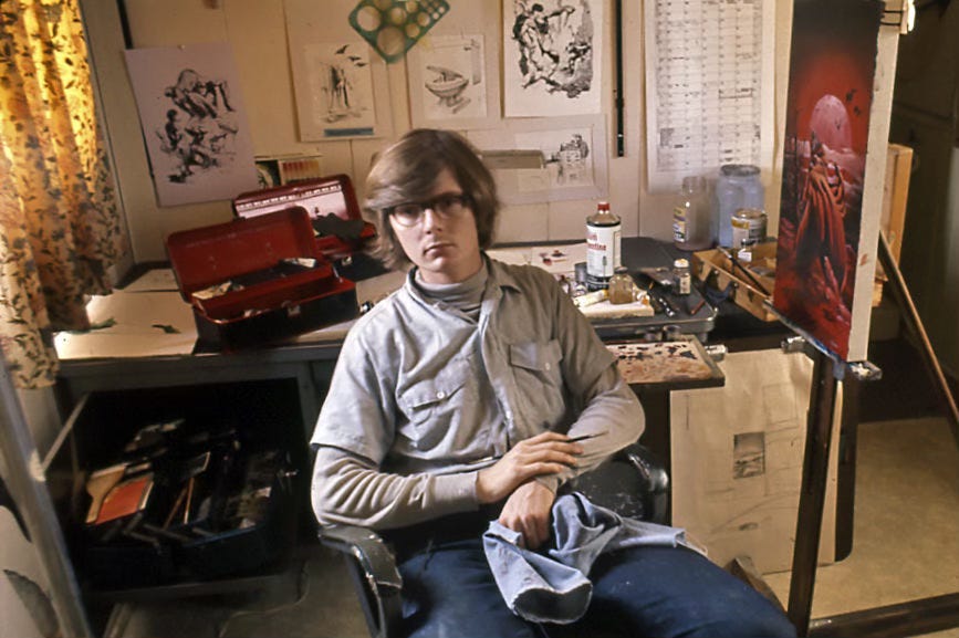 Michael Whelan photographed at age 20 in Cupertino, CA. Tacked on the board over his desk are references from various artists. He's seated with elbows on the arms of a desk chair with brush in one hand which rests on his opposite wrist. He holds a cloth over his lap. Next to him a vibrant red painting sits on his easel. Michael Whelan photographed at age 20 in Cupertino, CA. Tacked on the board over his desk are references from various artists. He's seated with elbows on the arms of a desk chair with brush in one hand which rests on his opposite wrist. He holds a cloth over his lap. Next to him a vibrant red painting sits on his easel.