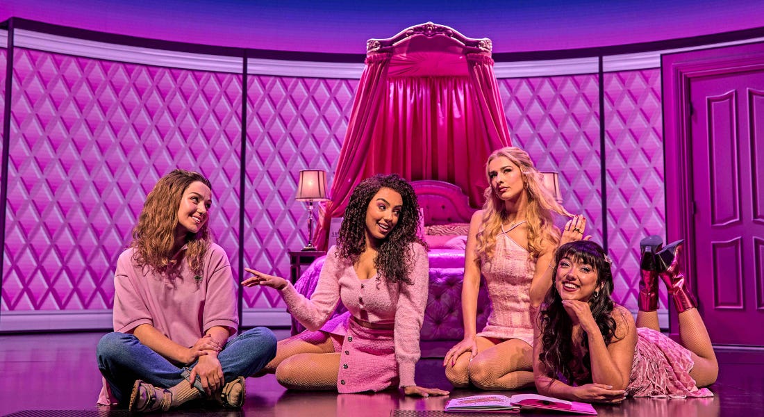 Four girls sat on a pink stage wearing pink outfits, with a pink bed behind them
