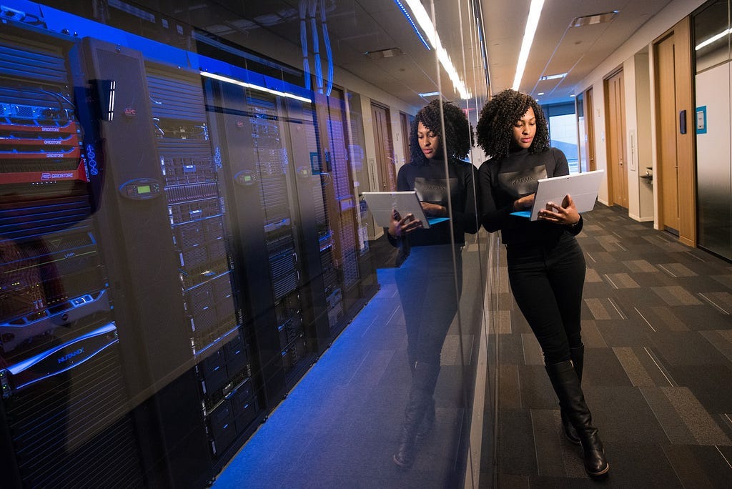 Woman in black top using Surface laptop as she stands beside large stacks of computer hardware.