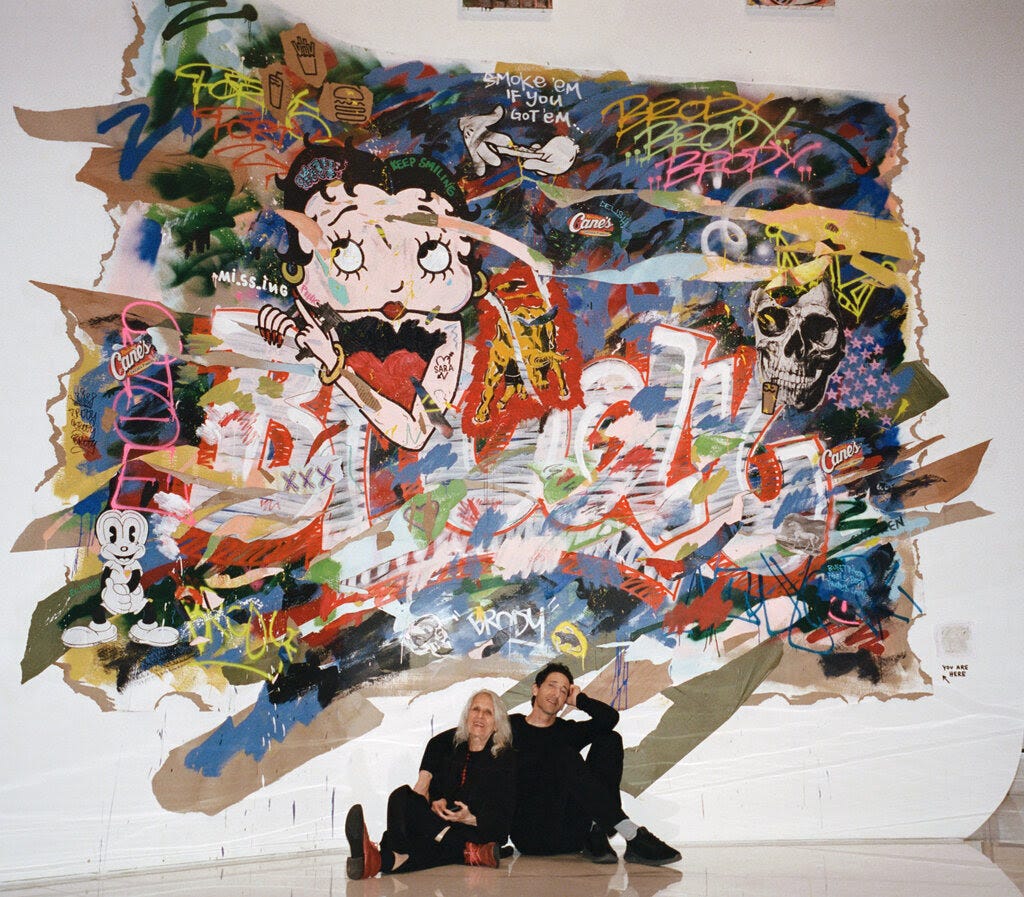 Adrien Brody and Sylvia Plachy sit on the floor of an art gallery, in front of a large collage that includes cartoon characters like Betty Boop.