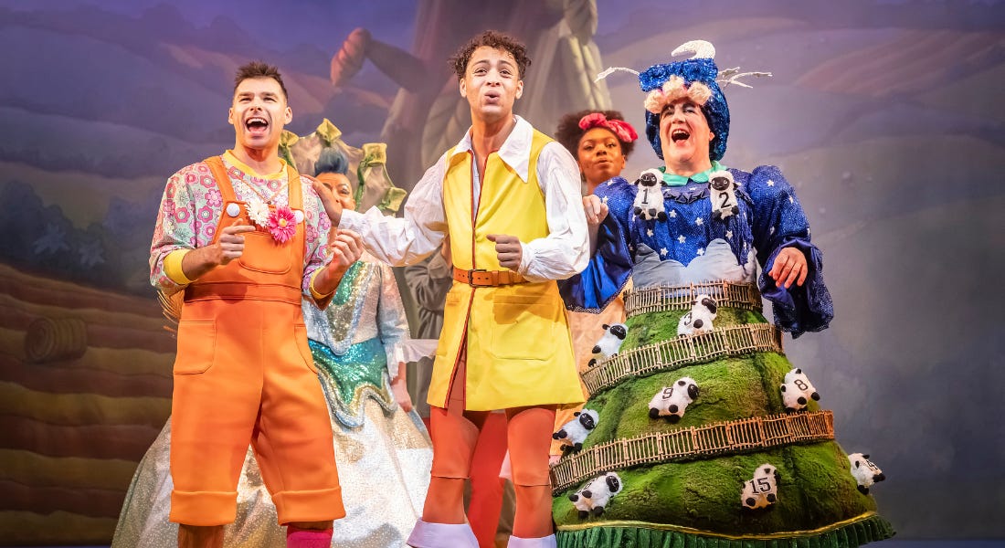 A group of five actors, all in brightly coloured costumes, stand in a huddle on the stage, with a giant beanstalk behind them. They are all singing, with smiles on their faces.
