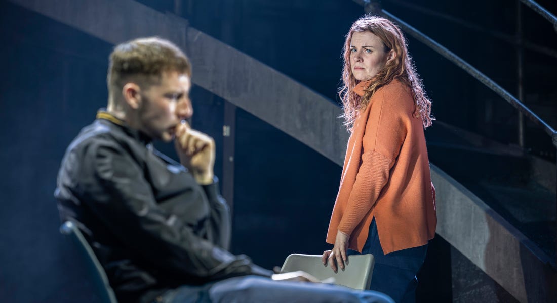 A man and woman sit in chairs beside each other. The man wears a brown cardigan and is speaking and using his hands to express himself. The woman, wearing a blue cardigan, looks at him with wide eyes and mouth open.