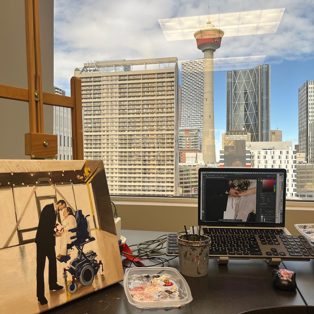 Athena's desk at the studio with the unfinished wedding painting on a small desktop easel and the reference image on her laptop. The window behind her desk shows downtown Calgary with office buildings and the Calgary Tower. Athena's desk at the studio with the unfinished wedding painting on a small desktop easel and the reference image on her laptop. The window behind her desk shows downtown Calgary with office buildings and the Calgary Tower.