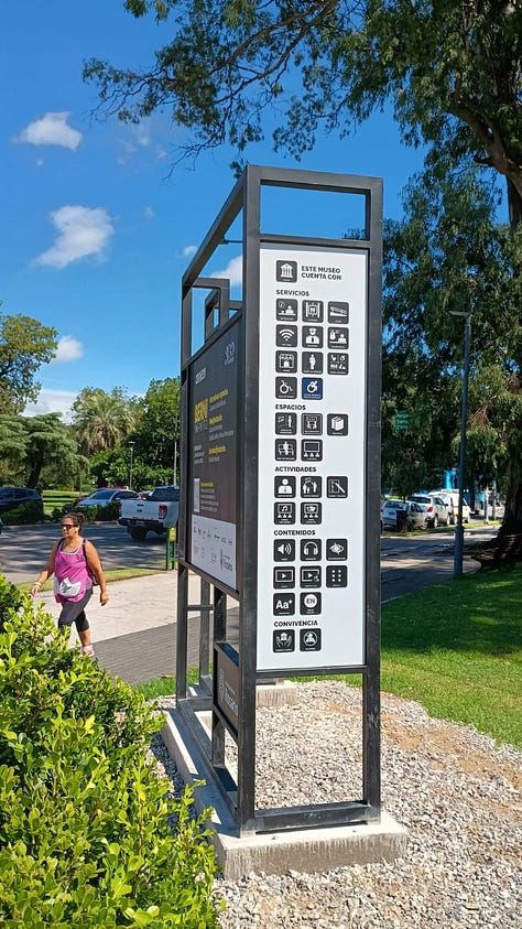 1- Fotografía de una cartelería externa de un museo, hecha de estructura de metal de aproximadamente unos tres metros de altura y sesenta centimetros de ancho. Se encuentra ubicado en el medio de un parque, en un sector donde hay tránsito peatonal. En la cartelería hay pictogramas que indican los espacios, servicios, contenidos y actividades de el museo castagnino 2- Fotografía de una cartel externa de un museo ubicado en el jardin previo al ingreso al edificio. El cartel es un rectángulo horizontal con un pie metalico que entra en el suelo. Da la bienvenida y expone pictogramas que indican los espacios, servicios, contenidos y actividades con los que cuenta el museo. 3- Fotografia de una pared en el interior de un museo. la pared esta pintada de negro y tiene en color blanco la leyenda con el nombre dle museo nacional del grabado y un texto. Al costado derecho hay un listado de pictogramas que indican los espacios, servicios, contenidos y actividades con los que cuenta el museo. 4- Fotografia de una pared en el interior de un museo. la pared es blanca y tiene en el centro un recuadro con borde negro y en su interior un texto que da la bienvenida. Debajo hay un listado de pictogramas que indican los espacios, servicios, contenidos y actividades con los que cuenta el museo. 5- Fotografia de un sector de una exhibición. En la pared hay cuatro imagenes enmarcadas y debajo en un exhibidor hay folios aillados con documentos antiguos y otras imagenes. En un costado se encuentra un pictograma y un código qr. 6-Fotografia de una sala de exhibición de un museo de arte contemporaneo. En el centro hay una mujer de pie agarrando el celular con las dos manos para sacar una fotografia a un dispositivo que hay en una de las paredes de la exhibición. detrás de ella, al final de la sala hay mas personas de pie y sentadas.