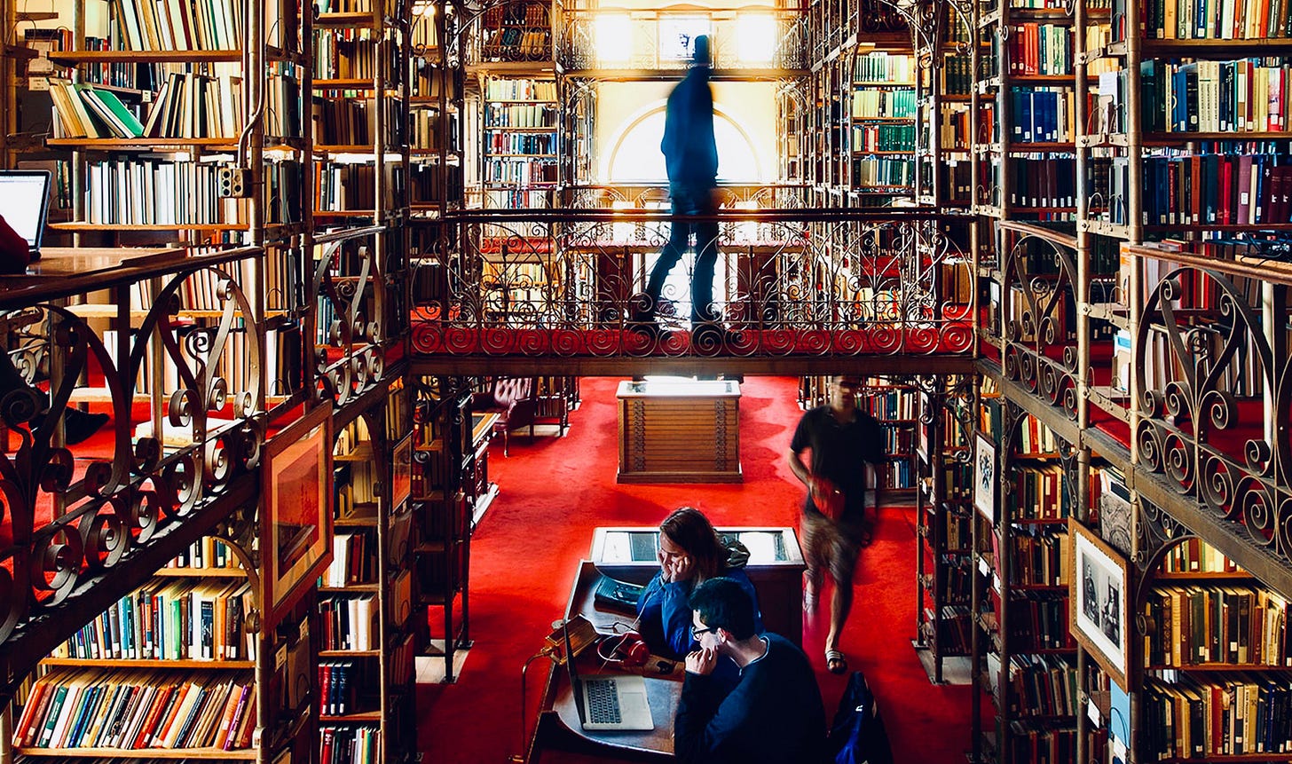 Photograph of stacks of books in an old-fashioned library. There are some people.