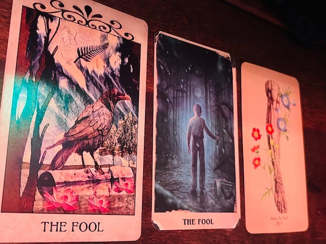 Three tarot cards labeled “The Fool” are laid side by side on a wooden surface. Each card features a different artistic style, including a bird illustration, a lone figure in a forest, and a minimalist design with flowers.