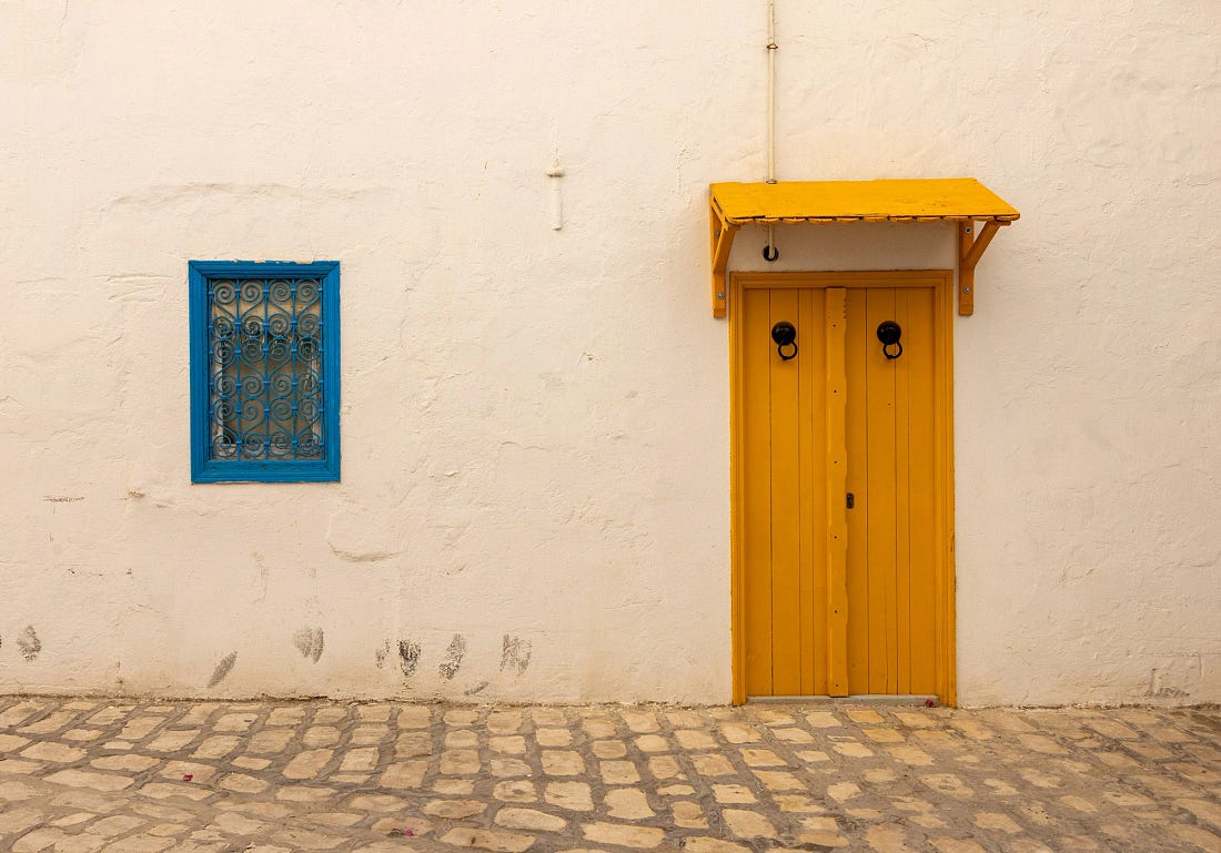 Mustard-yellow wooden door and blue iron-grilled window set into a whitewashed wall in the Tunis Medina, with a stone-paved street in the foreground.