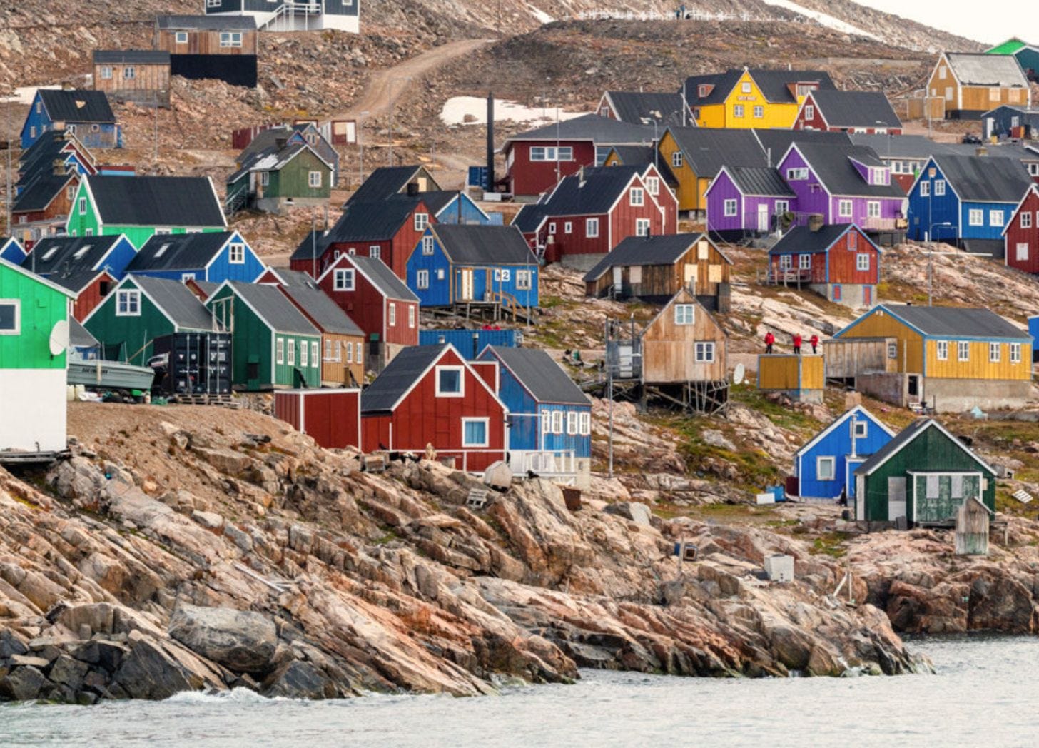 Wooden houses painted in bright colors including red, purple, green, dark blue, and yellow, built on stark rocky terrain. Each house has a pitched black or dark roof. The homes sit at various levels on the rocks. Patches of grass grow between them. Water in the foreground.