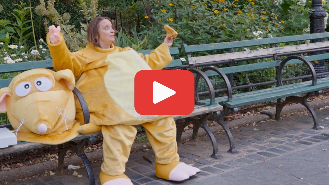 Photo of Rachel Dratch dressed in a rat suit and eating pizza on a bench in a park