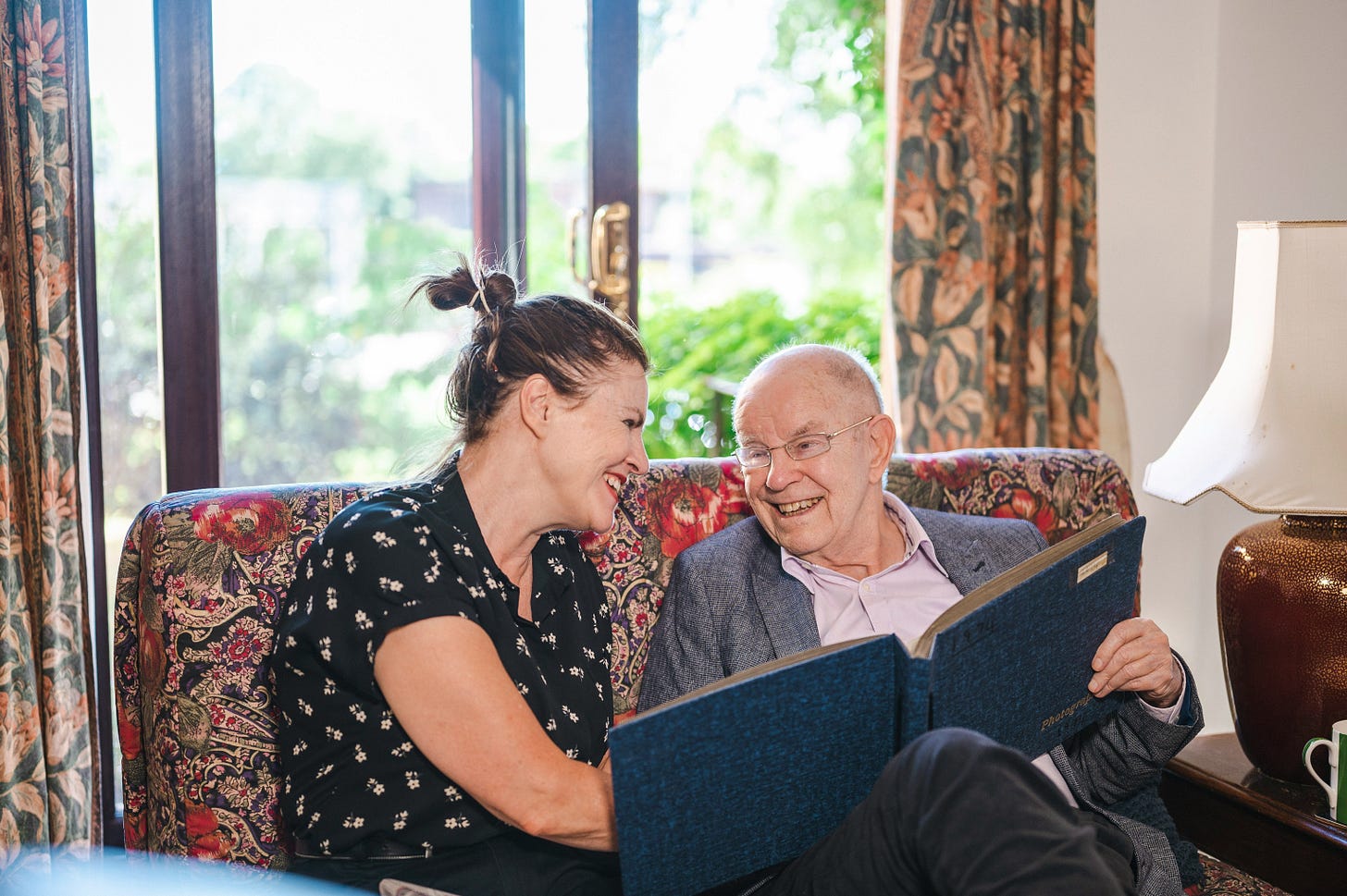 a woman in a ponytail in her 30s sitting on an antique couch with an elderly bald man looking at a photo album together and laughing a woman in a ponytail in her 30s sitting on an antique couch with an elderly bald man looking at a photo album together and laughing