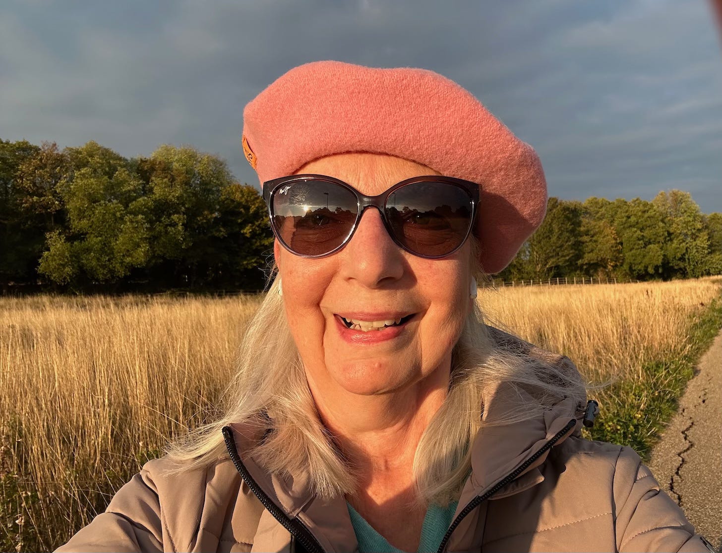 head and shoulders image of a woman smiling wearing sunglasses a coat and a pink beret walking towards the sun with a field and trees in the background