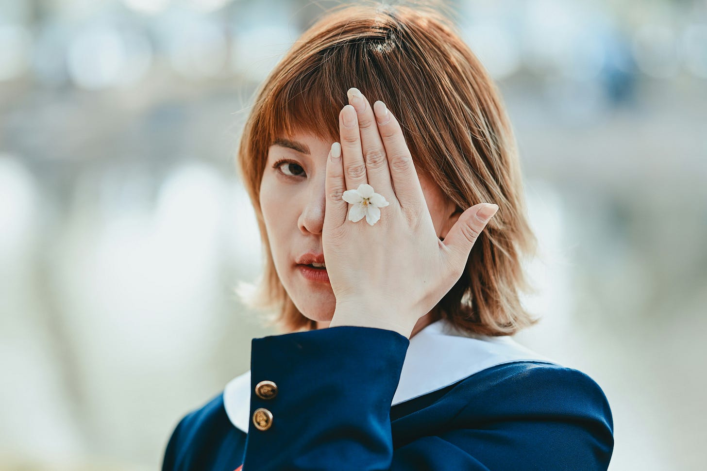 A young woman in a dark blue uniform holds her hand over one eye, a small white flower resting on her palm, her other eye looking directly at the camera, evoking the partial concealment at the heart of the authentic creative process and what we choose to hide versus reveal.