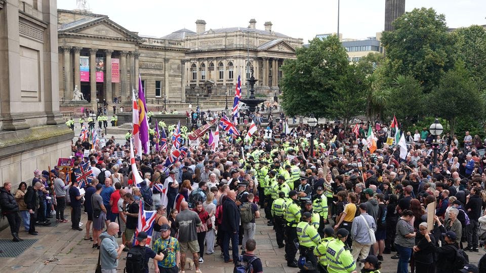 People hold Union Jack flags and colorful banners. Police officers in high-visibility jackets form a barrier between groups. 