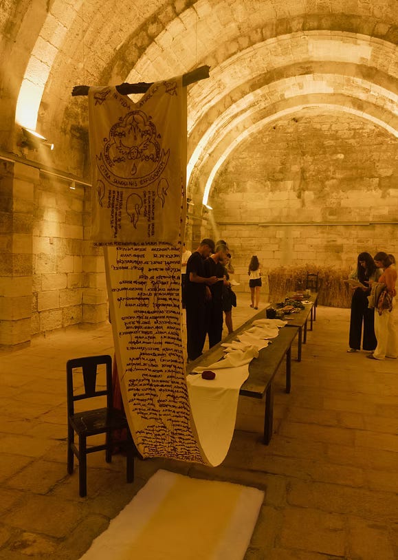 Sala in pietra con volte illuminate; al centro un lungo tavolo e un grande drappo verticale ricamato, osservati da alcuni visitatori. Sala in pietra con volte illuminate; al centro un lungo tavolo e un grande drappo verticale ricamato, osservati da alcuni visitatori.