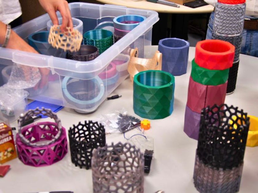 Diversos recipientes cilíndricos produzidos em impressão 3D sobre uma mesa branca. Os objetos possuem texturas variadas, como tramas vazadas, padrões geométricos e colmeias, em cores como verde, roxo, azul, rosa e preto.