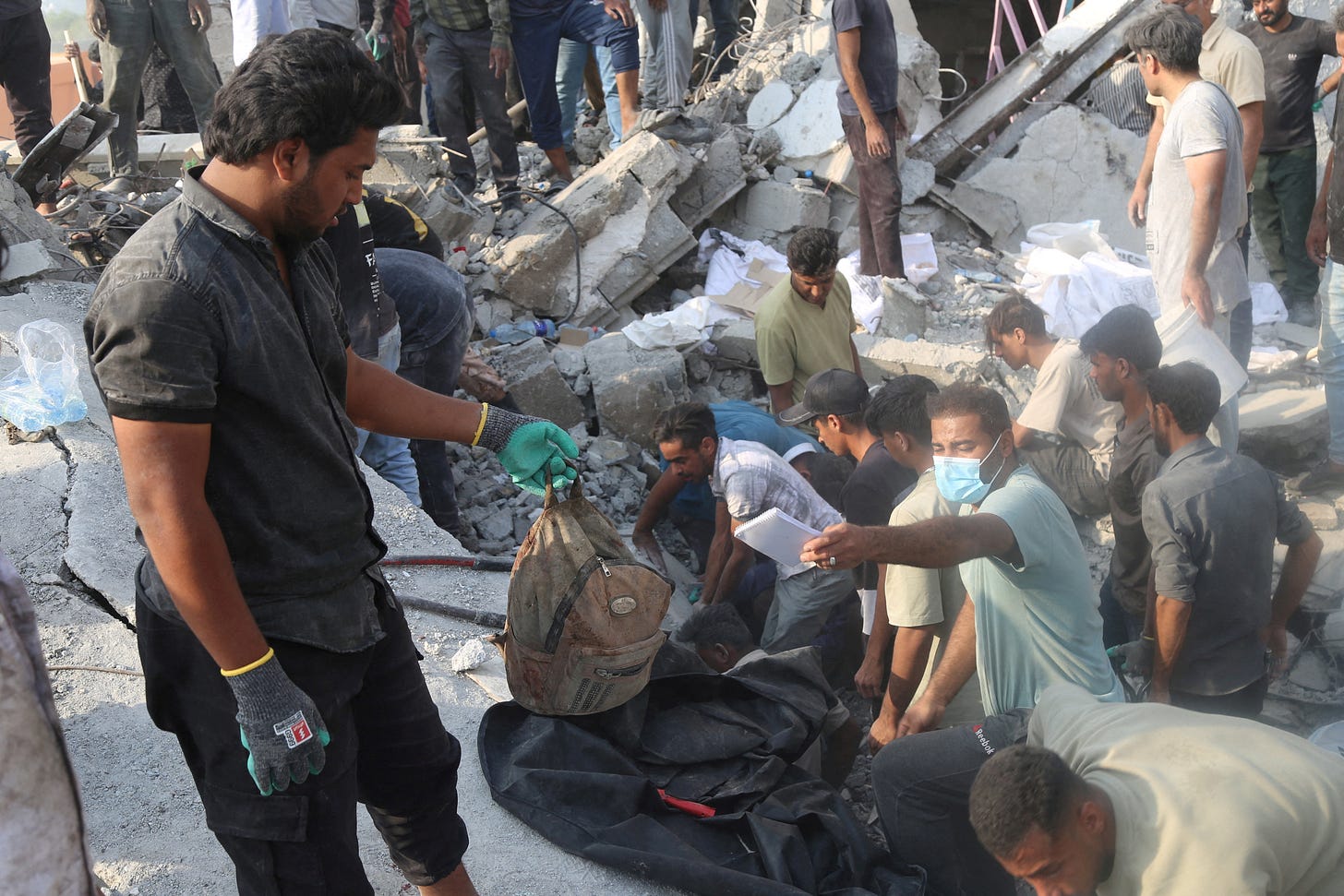 Rescuers and bystanders search through the rubble at the site Iranian authorities identified as the Shajareh Tayyebeh girls’ school in Minab, southern Iran. In the foreground, a man in gloves holds up a dust-covered child’s backpack over a black body bag while others dig through broken concrete, twisted rebar, and scattered belongings in the collapsed building.