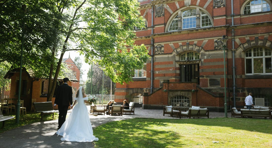 A bride and a groom walking towards a large brick pumping house on a winding path A bride and a groom walking towards a large brick pumping house on a winding path