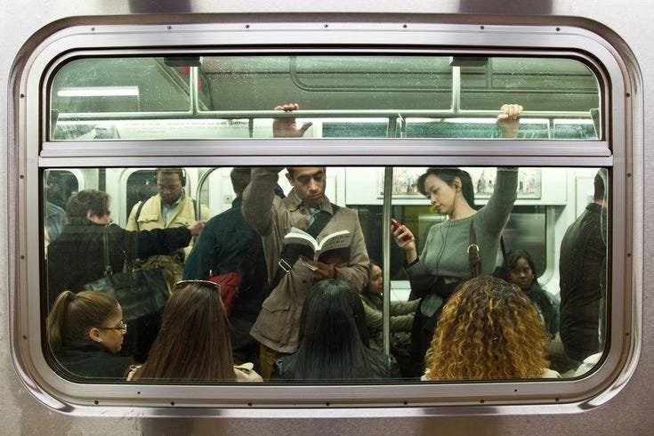 This may contain: people standing on a subway train looking at their phones and reading books while others look out the window