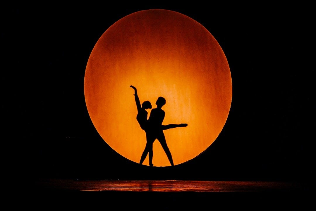 Dancers silhouetted against a giant, orange orb. Dancers silhouetted against a giant, orange orb.