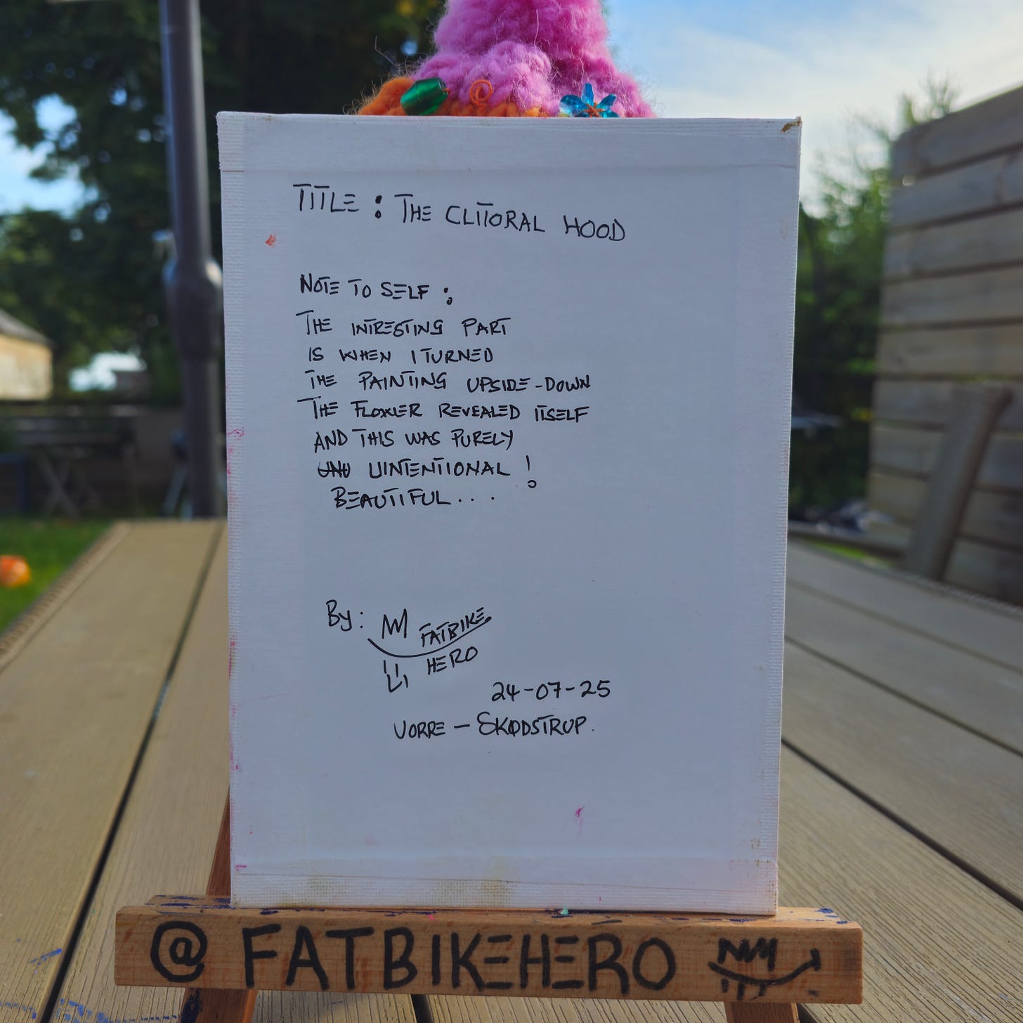 Alt Text: The back of a white canvas with handwritten black text. It reads: "TITLE: THE CLITORAL HOOD. NOTE TO SELF: THE INTERESTING PART IS WHEN I TURNED THE PAINTING UPSIDE-DOWN THE FLOWER REVEALED ITSELF AND THIS WAS PURELY UNINTENTIONAL! BEAUTIFUL..." Signed by M / Fatbike Hero, dated 24-07-25, Vorre-Skødstrup. Alt Text: The back of a white canvas with handwritten black text. It reads: "TITLE: THE CLITORAL HOOD. NOTE TO SELF: THE INTERESTING PART IS WHEN I TURNED THE PAINTING UPSIDE-DOWN THE FLOWER REVEALED ITSELF AND THIS WAS PURELY UNINTENTIONAL! BEAUTIFUL..." Signed by M / Fatbike Hero, dated 24-07-25, Vorre-Skødstrup.