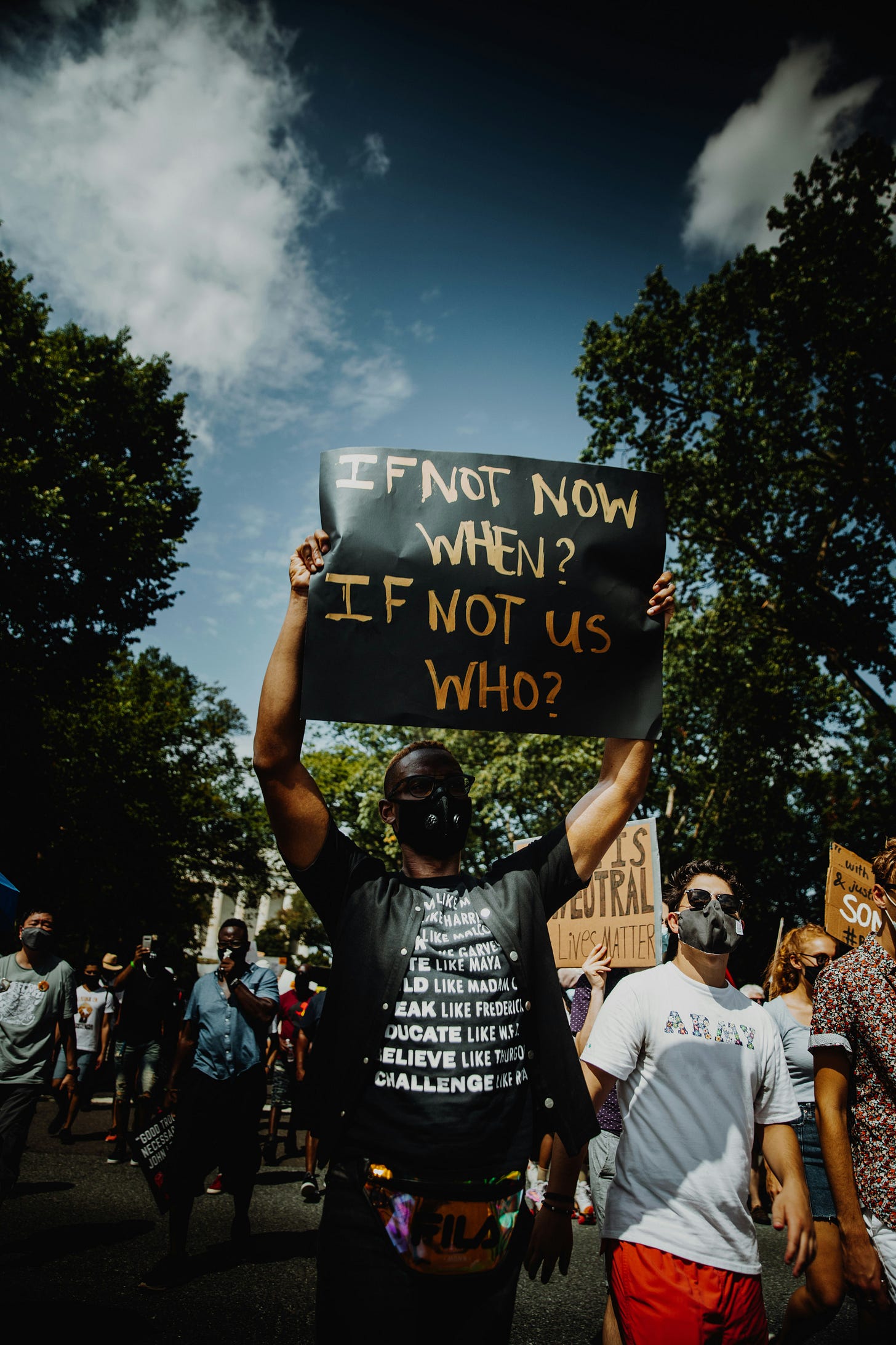 A Black protester wearing a face mask holds a sign reading “If not now, when? If not us, who?” while marching alongside a diverse crowd of demonstrators on a tree-lined street under a blue sky.