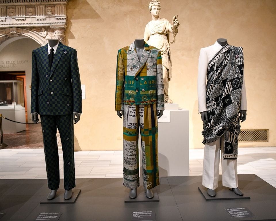 Three intricately tailored menswear ensembles displayed at The Met's Costume Institute exhibit on Black style. The looks celebrate superfine tailoring and cultural expression: a dark green checkerboard suit, a vibrant printed suit covered in text referencing African diasporic narratives, and an off-white suit with a black-and-white woven sash resembling traditional African textiles. Positioned in front of a classical sculpture, the display highlights the power, precision, and elegance of Black sartorial excellence. Three intricately tailored menswear ensembles displayed at The Met's Costume Institute exhibit on Black style. The looks celebrate superfine tailoring and cultural expression: a dark green checkerboard suit, a vibrant printed suit covered in text referencing African diasporic narratives, and an off-white suit with a black-and-white woven sash resembling traditional African textiles. Positioned in front of a classical sculpture, the display highlights the power, precision, and elegance of Black sartorial excellence.