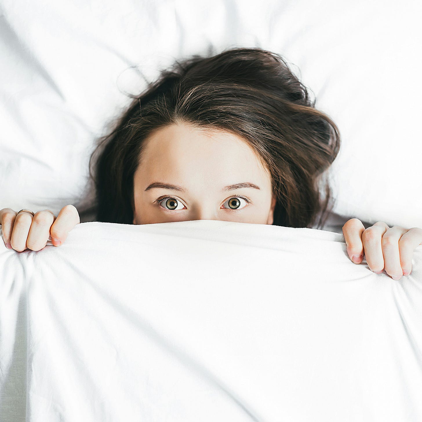 A person, female in appearance, is looking at the camera. The bottom half of their face is covered by a white blanket. Their eyes are wide as if in fear. A person, female in appearance, is looking at the camera. The bottom half of their face is covered by a white blanket. Their eyes are wide as if in fear.