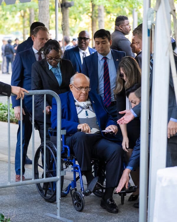 Rudolph W. Giuliani, exalcalde de Nueva York, lleva un traje de chaqueta y pantalones azul oscuro mientras está sentado en una silla de ruedas. Lleva un dispositivo médico en el torso. Lo rodean personas vestidas con trajes de trabajo.