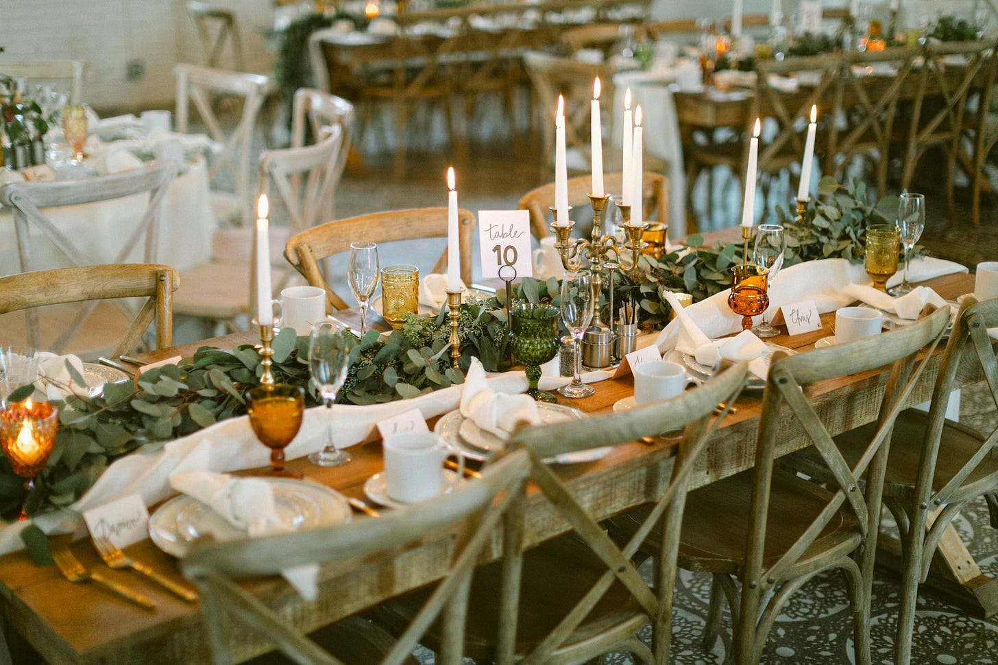 A long wooden table set for a gathering, lined with simple wooden chairs and decorated with greenery, folded white napkins, and place settings. Tall white candles in brass holders glow warmly down the center, while glassware and handwritten place cards add a personal, welcoming touch.