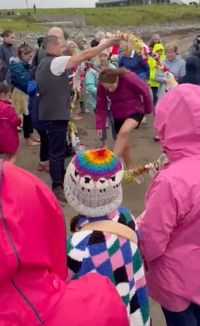 Russell holding up a braided willow belt wrapped in grasses and wishing scarves as a woman walks through it with people looking on Russell holding up a braided willow belt wrapped in grasses and wishing scarves as a woman walks through it with people looking on